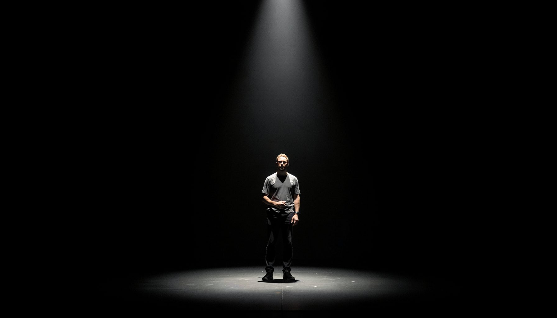 A lone actor on a bare stage, single harsh spotlight from above, deep black background, stark contrast
