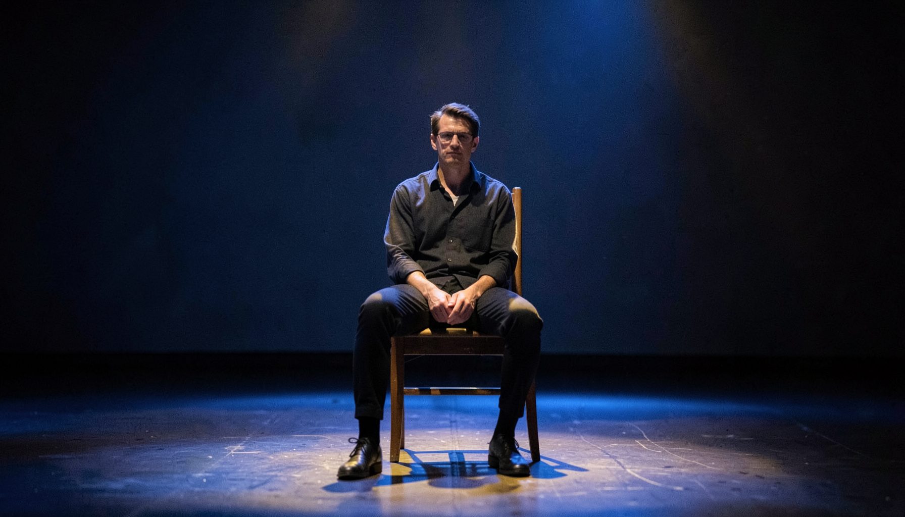 A lone performer seated on a simple wooden chair at centre stage, soft blue and gold lighting, quiet and reflective atmosphere
