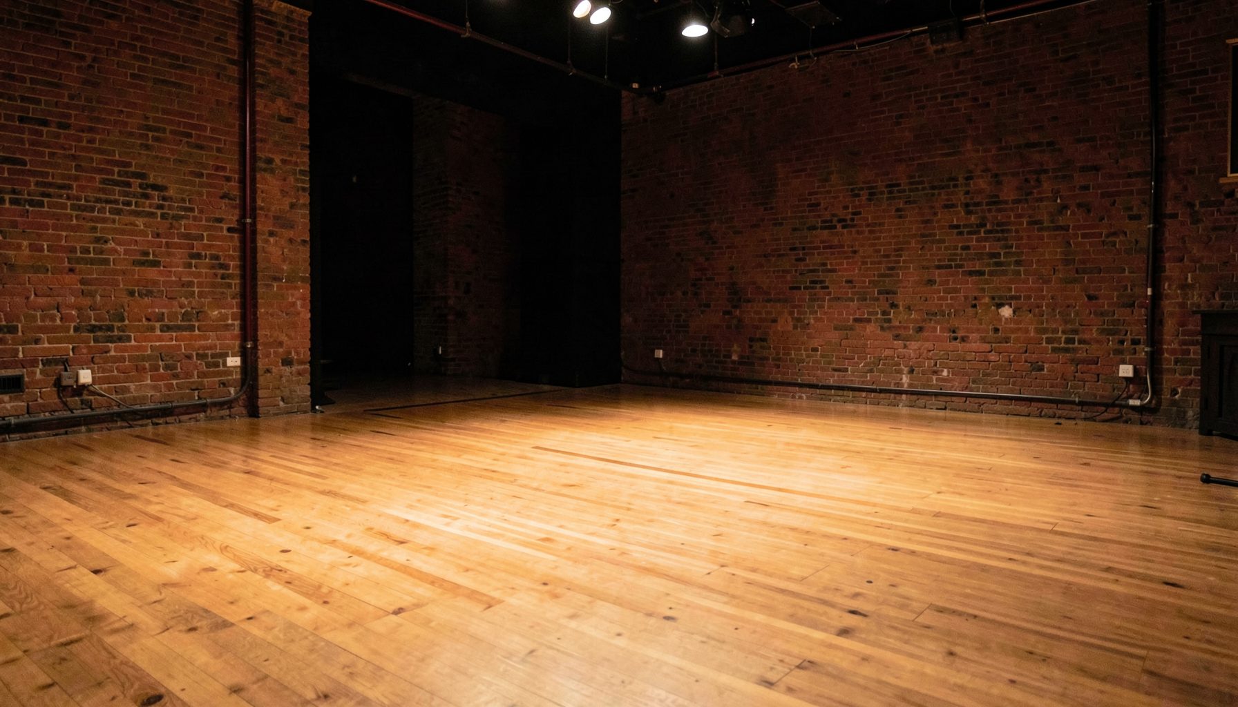 A warm, softly lit theatre rehearsal room with wooden floors and exposed brick walls, empty stage area in the background