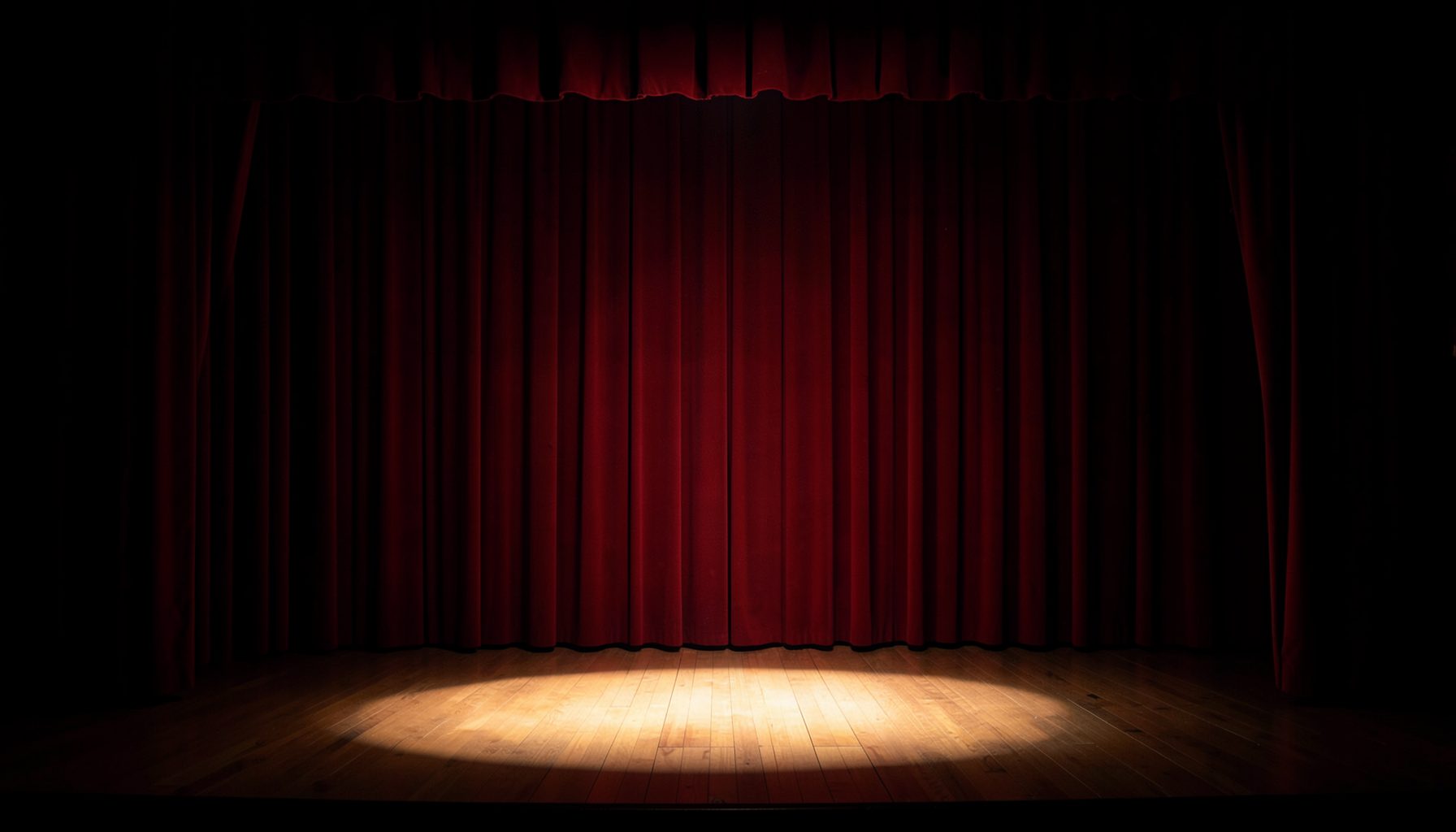 A warmly lit theatre stage with deep red curtains drawn back, a single spotlight pooling on the wooden floor, deep navy shadows around the edges