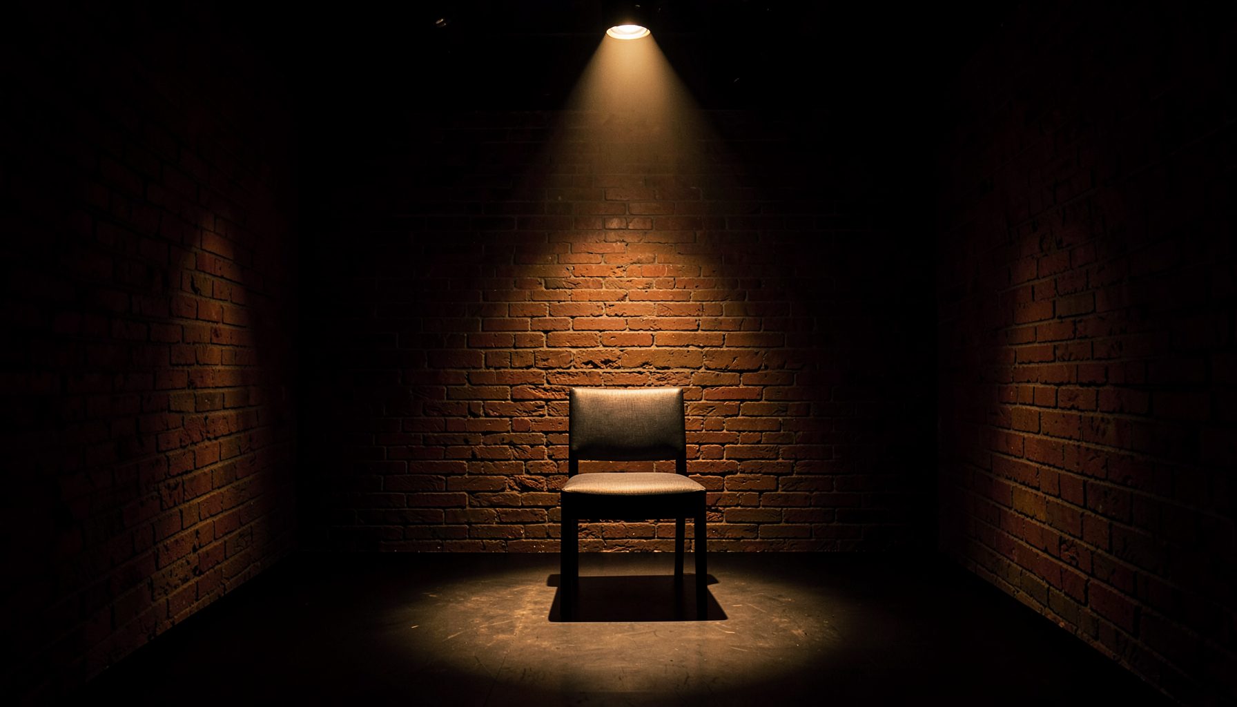 An intimate black-box theatre with exposed brick walls, a single chair under a spotlight, warm amber and deep shadow