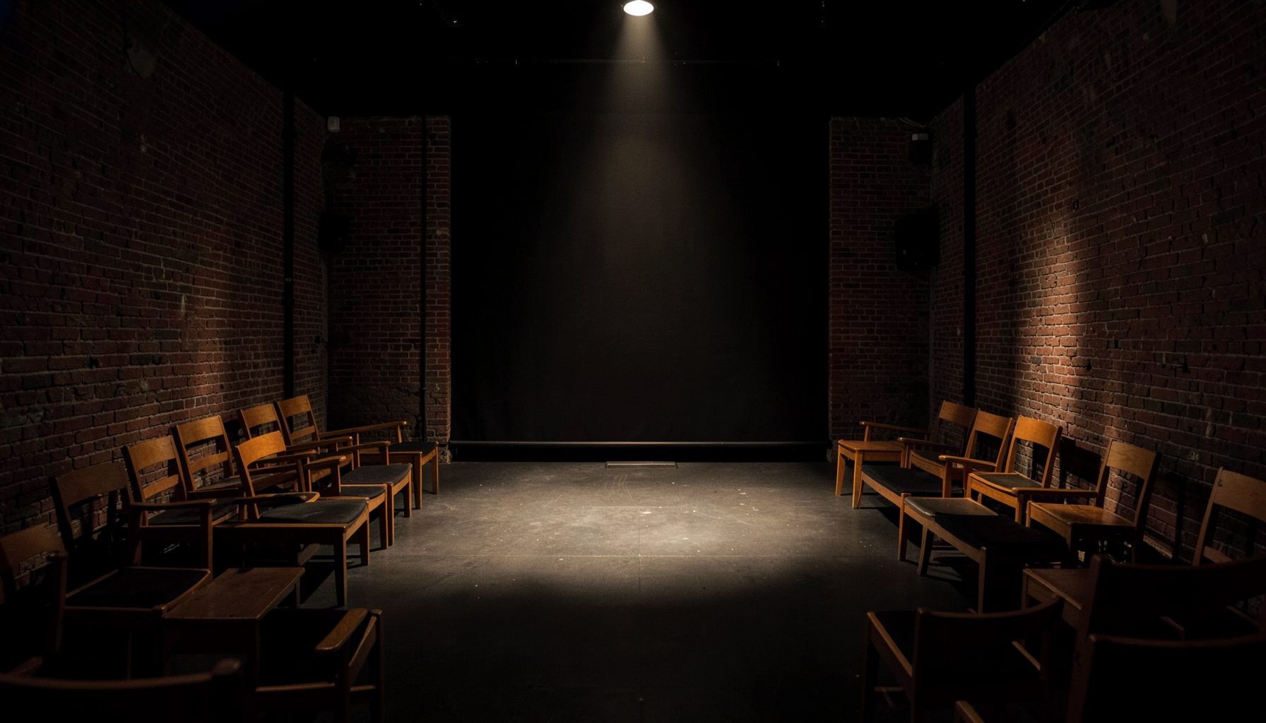 Intimate black-box theatre space with exposed brick walls and simple wooden seating, warm spotlight on an empty stage