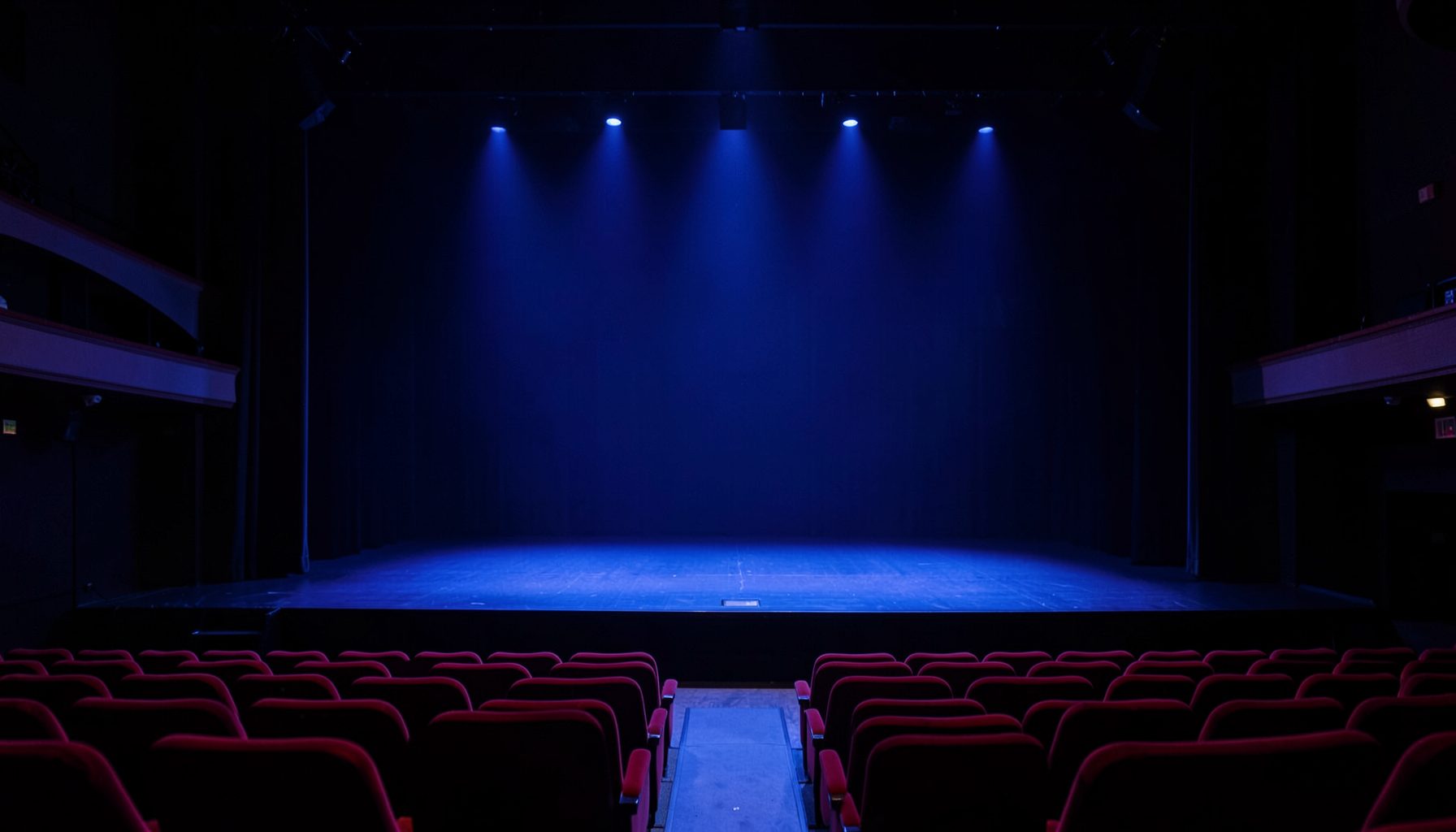 Wide theatre stage bathed in deep indigo and soft sky-blue stage lighting, empty red velvet seats in the foreground